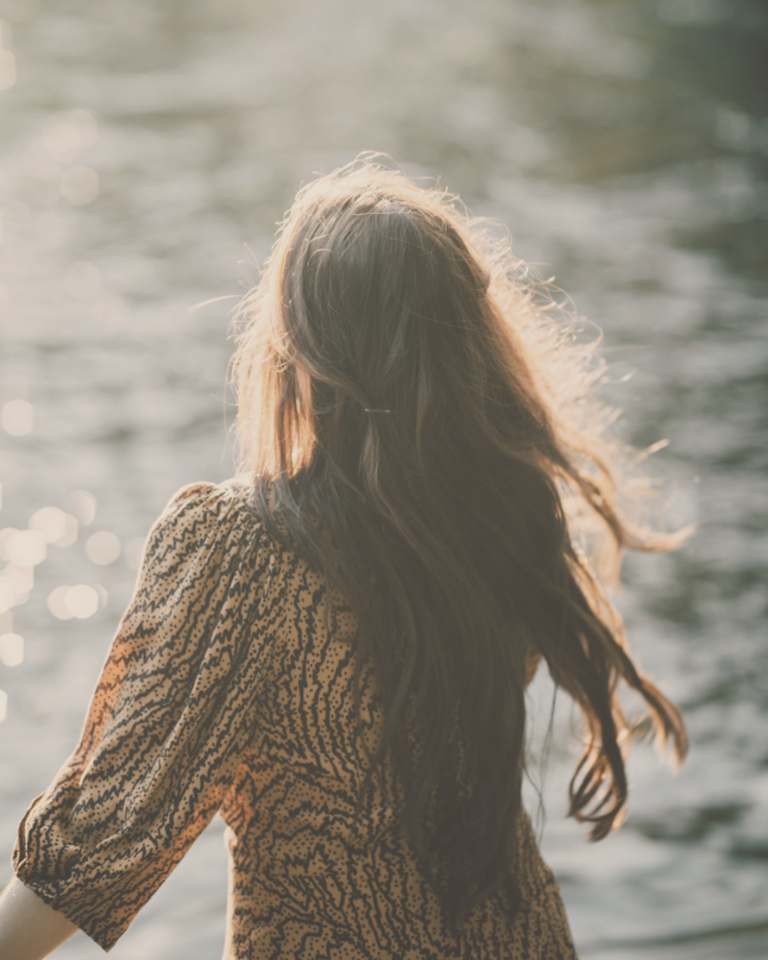 the back of a woman with beautiful floating brown hair looking at the reflection of the sun on the river immersed into the golden light of the sun