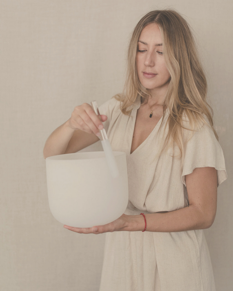 blonde woman wearing beige cotton dress holding a crystal singing bowl in her hand